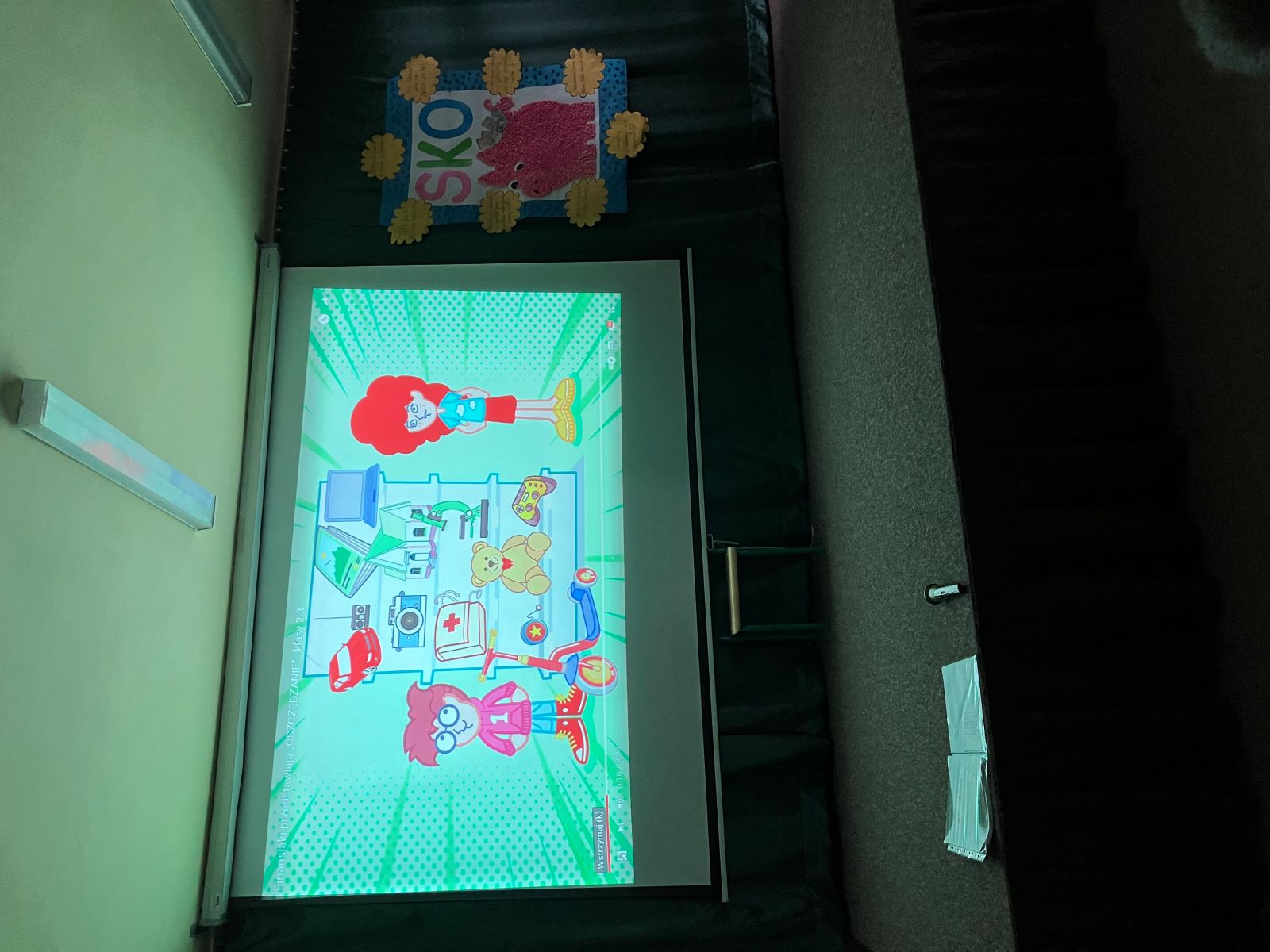 Scena, na której jest wyświetlany film edukacyjny, który przedstawia dwoje dzieci w sklepie z zabawkami. Obok zawieszona jest plansza z napisem SKO, dużą świnką-skarbonką, wokół której naklejone są hasła związane z oszczędzaniem