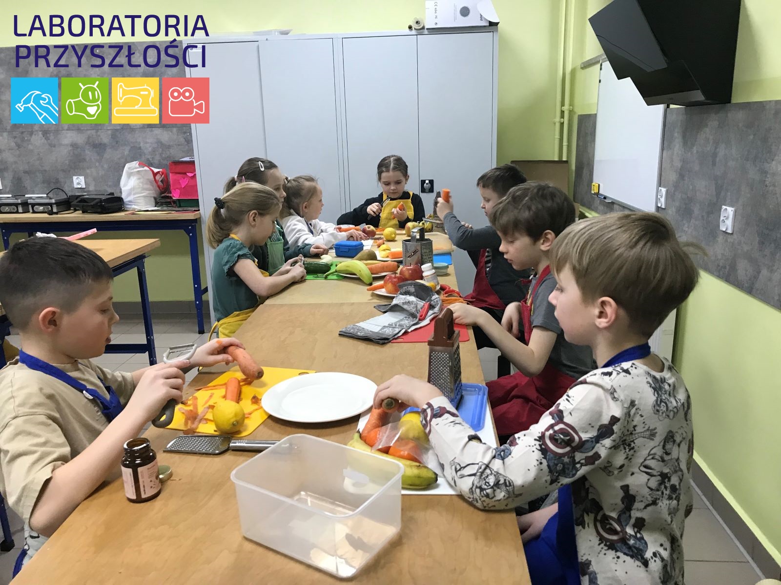 Na zdjęciu grupa uczniów  siedzi przy stole, przygotowują warzywa  i owoce na sałatkę. Na stole rozłożone deski do krojenia, talerze, tarki.