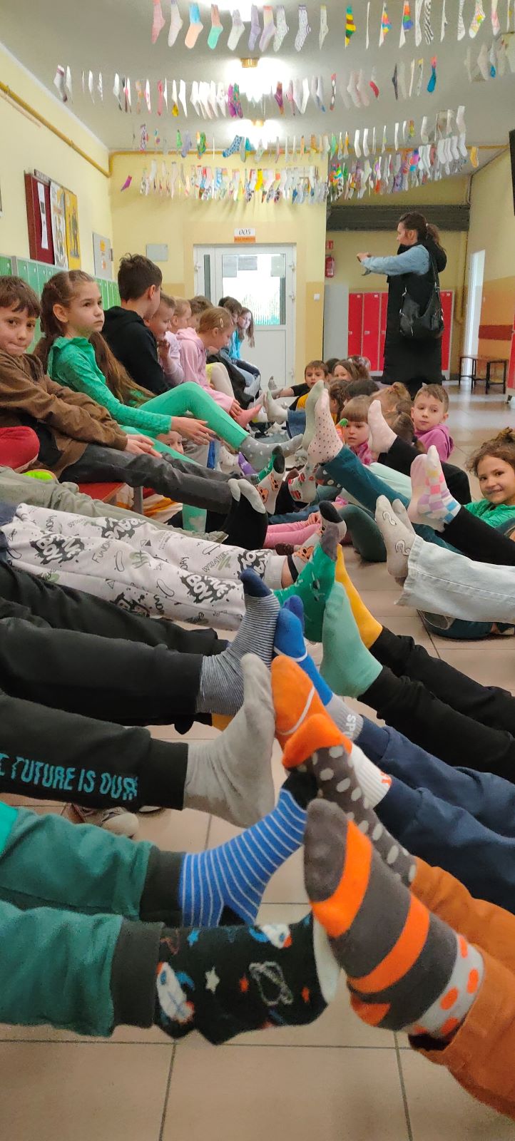 Uczniowie klas 1-3 siedząc na podłodze podnoszą nogi do góry pokazując założone kolorowe skarpetki nie do pary.