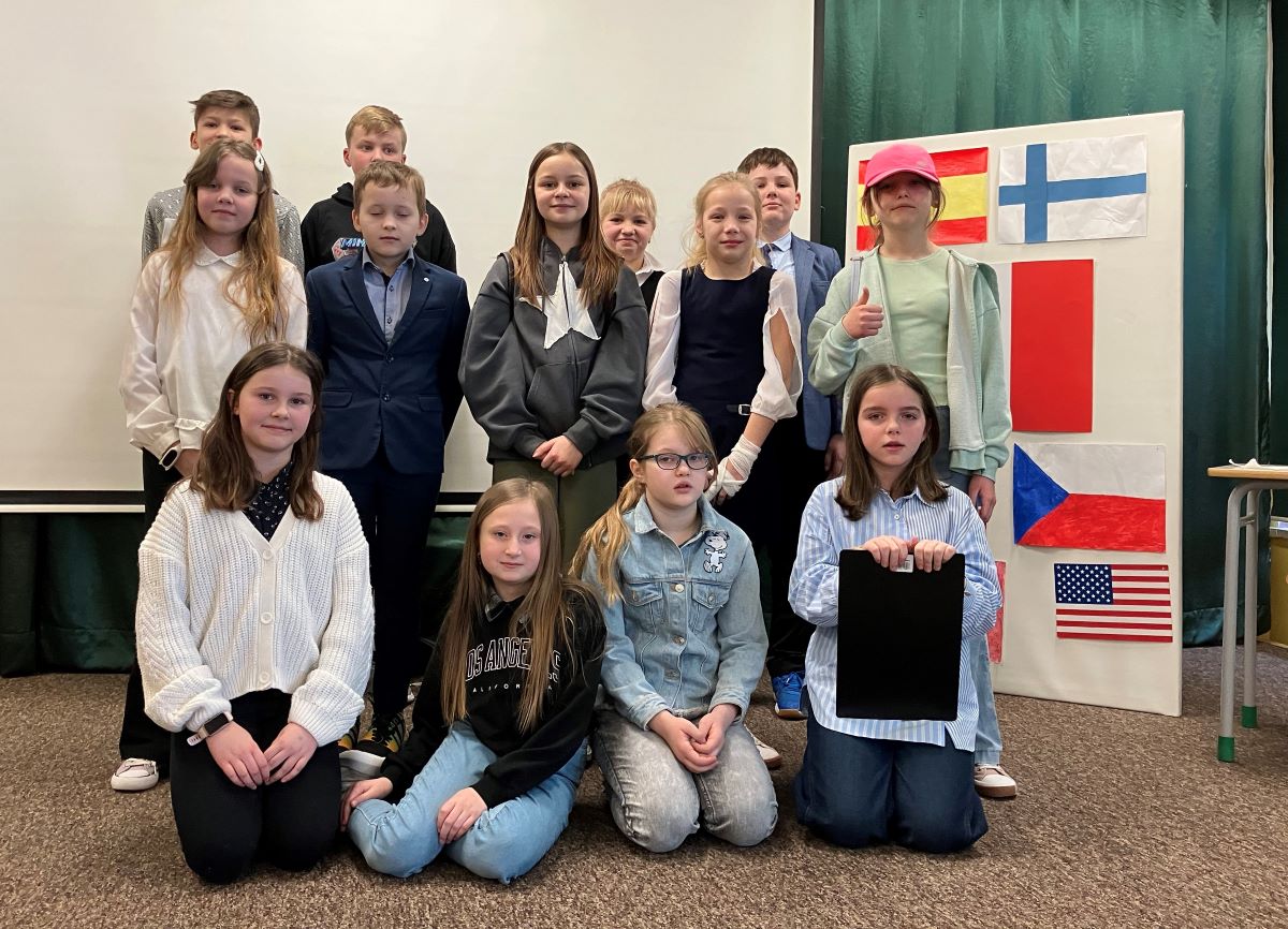 Grupa 13 uczniów, którzy brali udział w apelu. 4 chłopców i 9 dziewcząt na scenie, w tle ustawiona plansza z flagami różnych europejskich krajów.