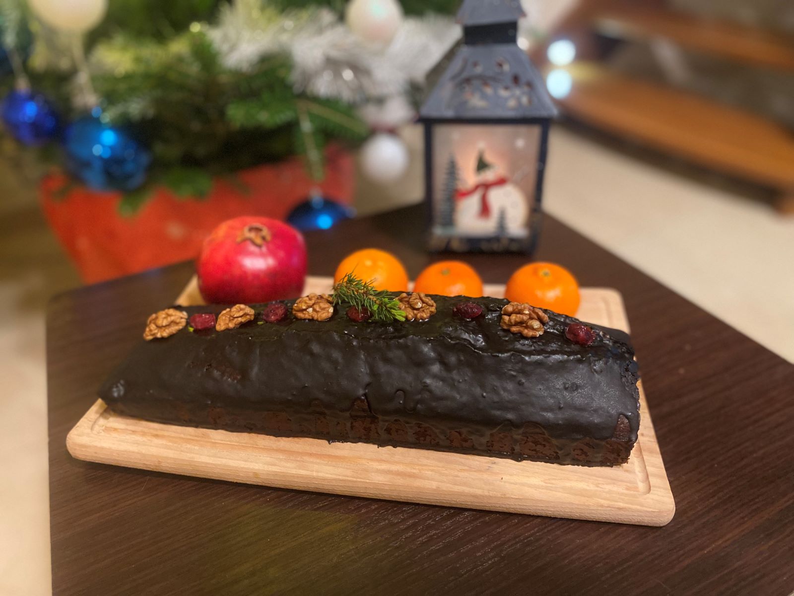 Na zdjęciu przedstawiony jest piernik z czekoladową polewą udekorowany orzechami laskowymi i żurawiną. W tle świąteczna dekoracja.