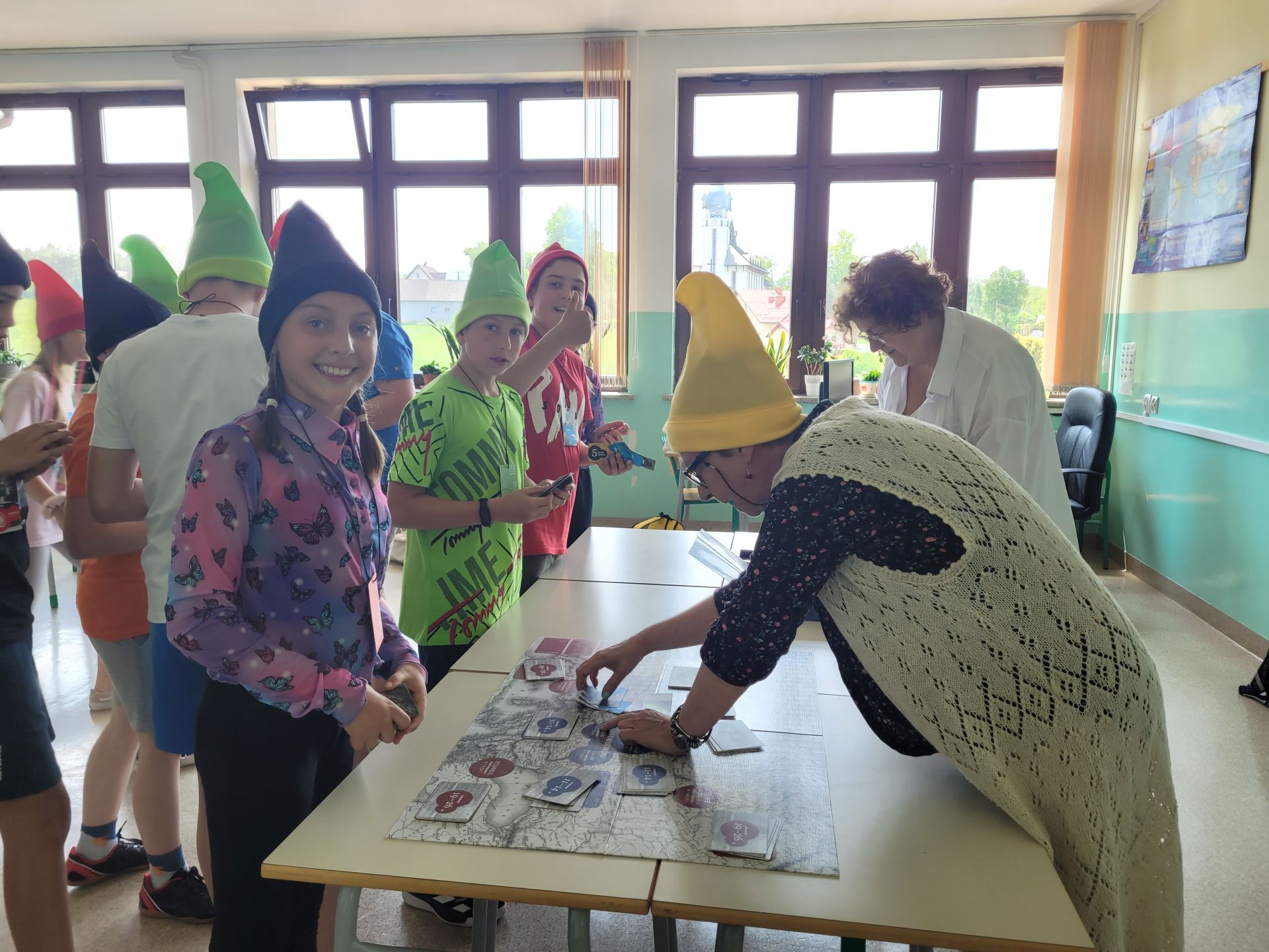 W sali lekcyjnej grupa dzieci gra w grę planszową ubrana w kolorowe, spiczaste czapeczki