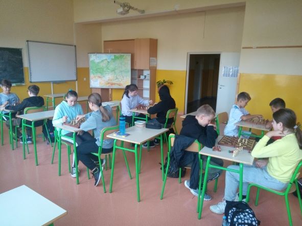 Dzieci w klasie siedzą przy stolikach na których znajdują się szachy Uczniowie grają w tą grę.