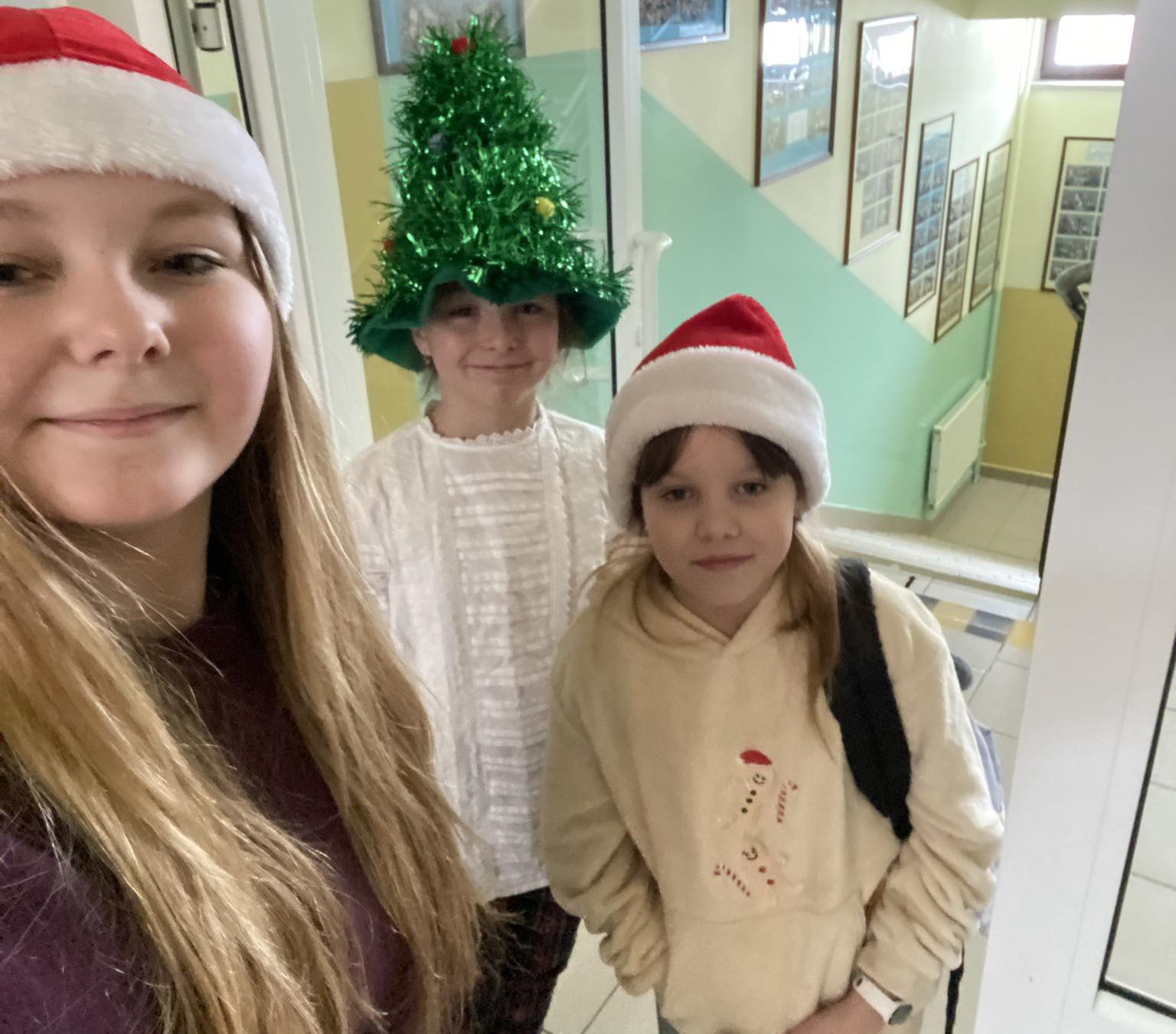 Na szkolny, korytarzu trzy dziewczynki pozują do zdjęcia. Dwie z nich ma ubrane czerwone czapki Mikołaja, dziewczynka pośrodku ma na głowie czapkę w kształcie dużej, zielonej choinki