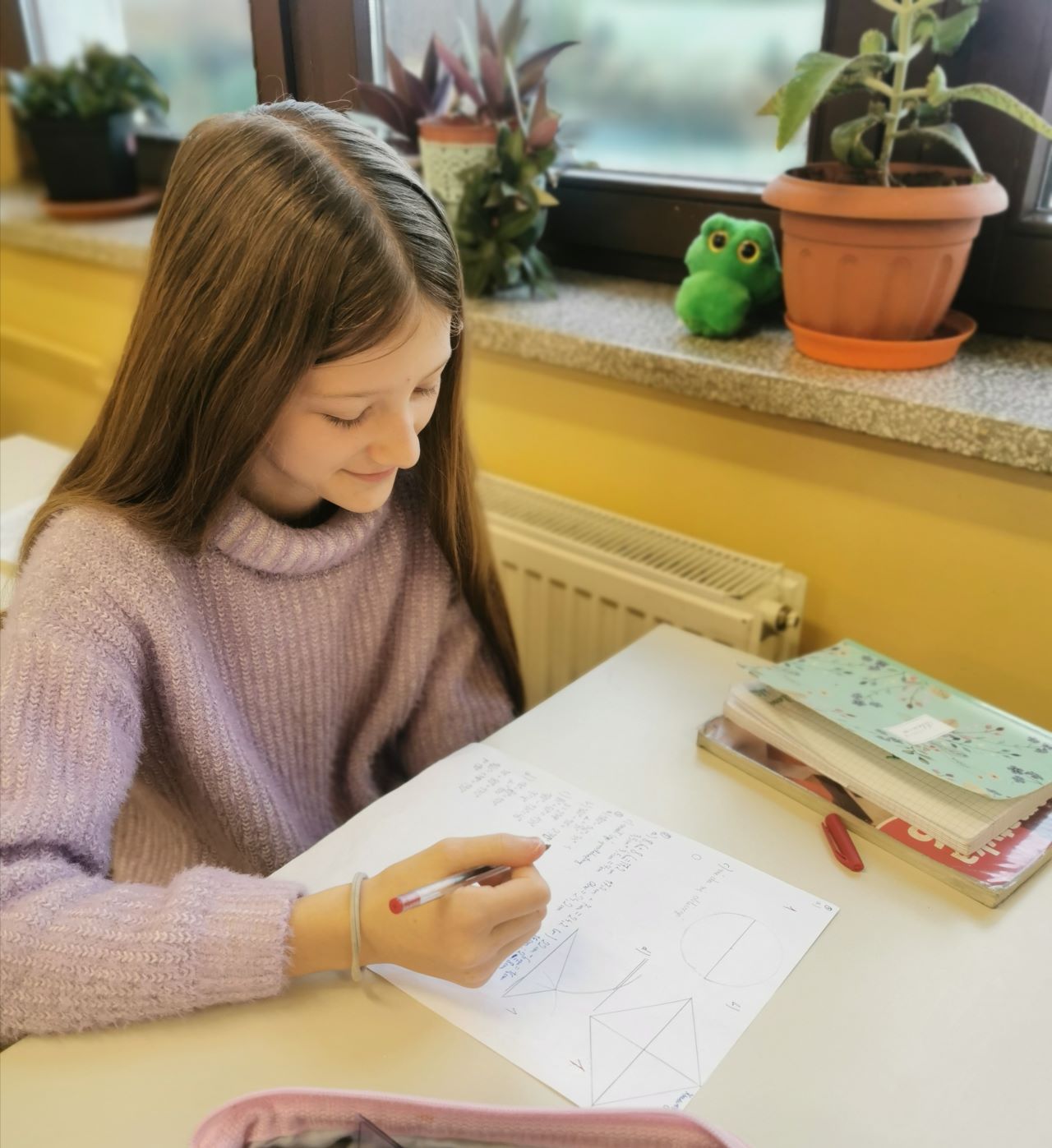 Na zdjęciu uczennica klasy szóstej siedzi w ławce , trzyma w ręce czerwony długopis i ocenia zestaw zadań z geometrii