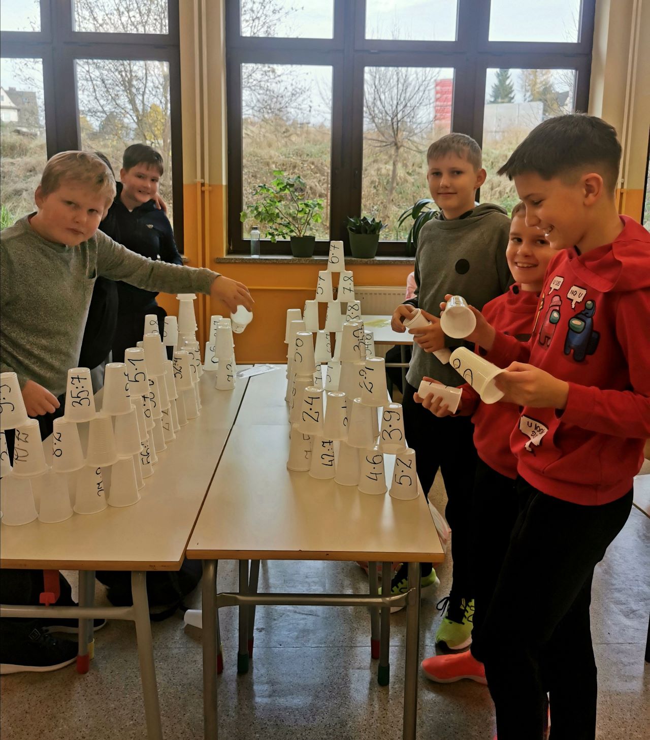 Pięcioro uczniów klasy piątej stoi przy stolikach  i układa wieże z plastikowych kubeczków. Na kubeczkach są napisane iloczyny lub ilorazy liczb