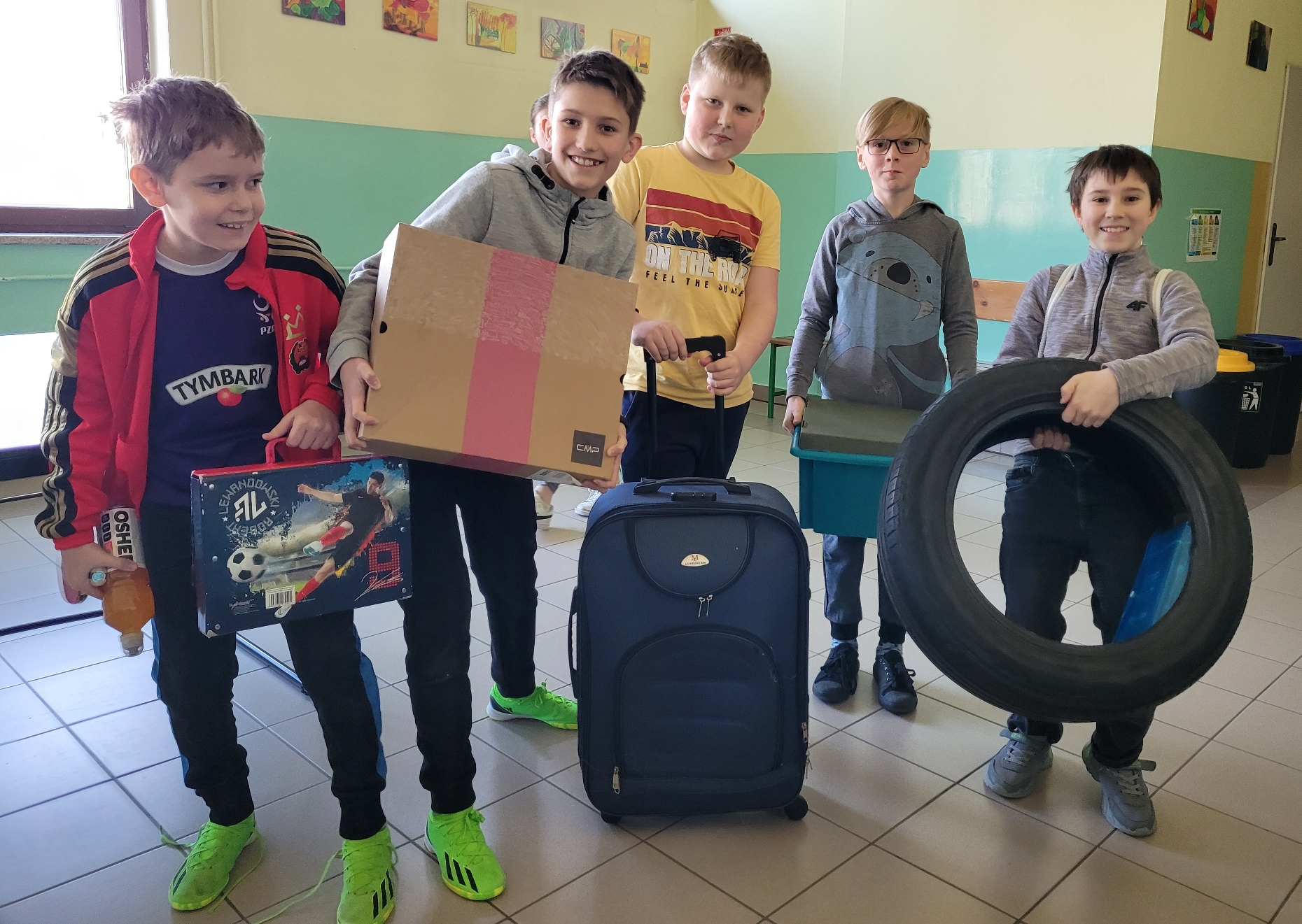 Grupa sześciu chłopców stoi z pudełkiem, walizką i oponą na szkolnym korytarzu.