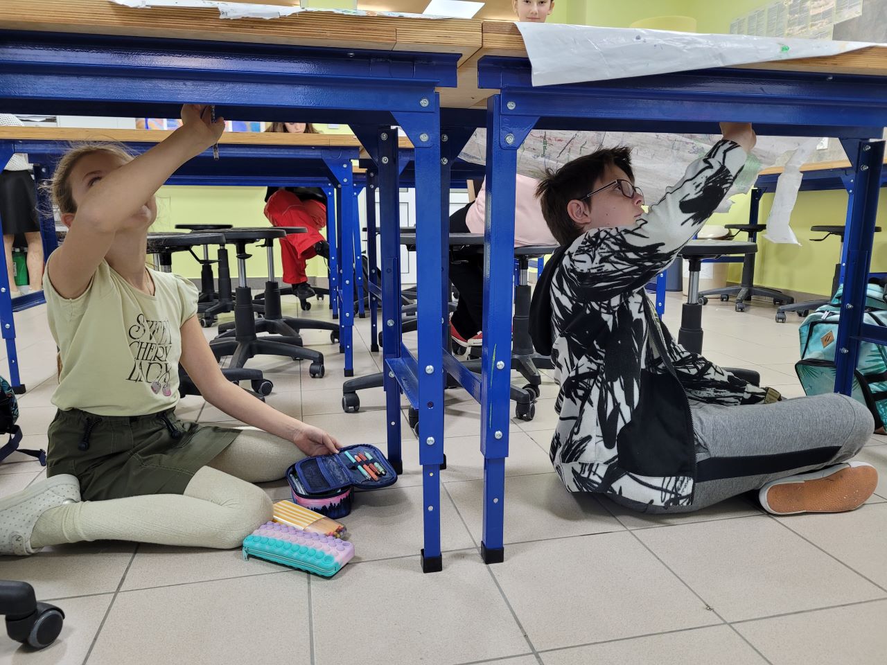 W sali lekcyjnej dwójka uczniów: uczennica i uczeń siedzą pod ławką z głową zadartą do góry i ręką uniesioną w stronę blatu