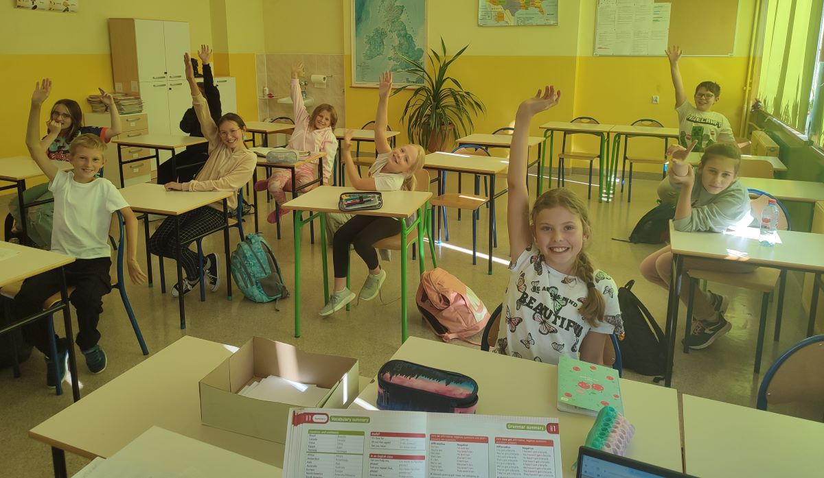 Uczniowie siedzący w klasie przy ławkach z podniesionymi rękami.