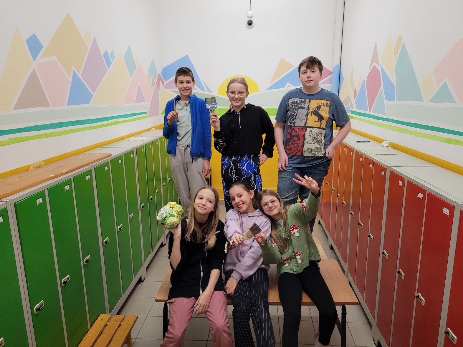 Na zdjęciu grupa uśmiechniętych dzieci na tle pomalowanej w różnobarwne kształty ścianie.