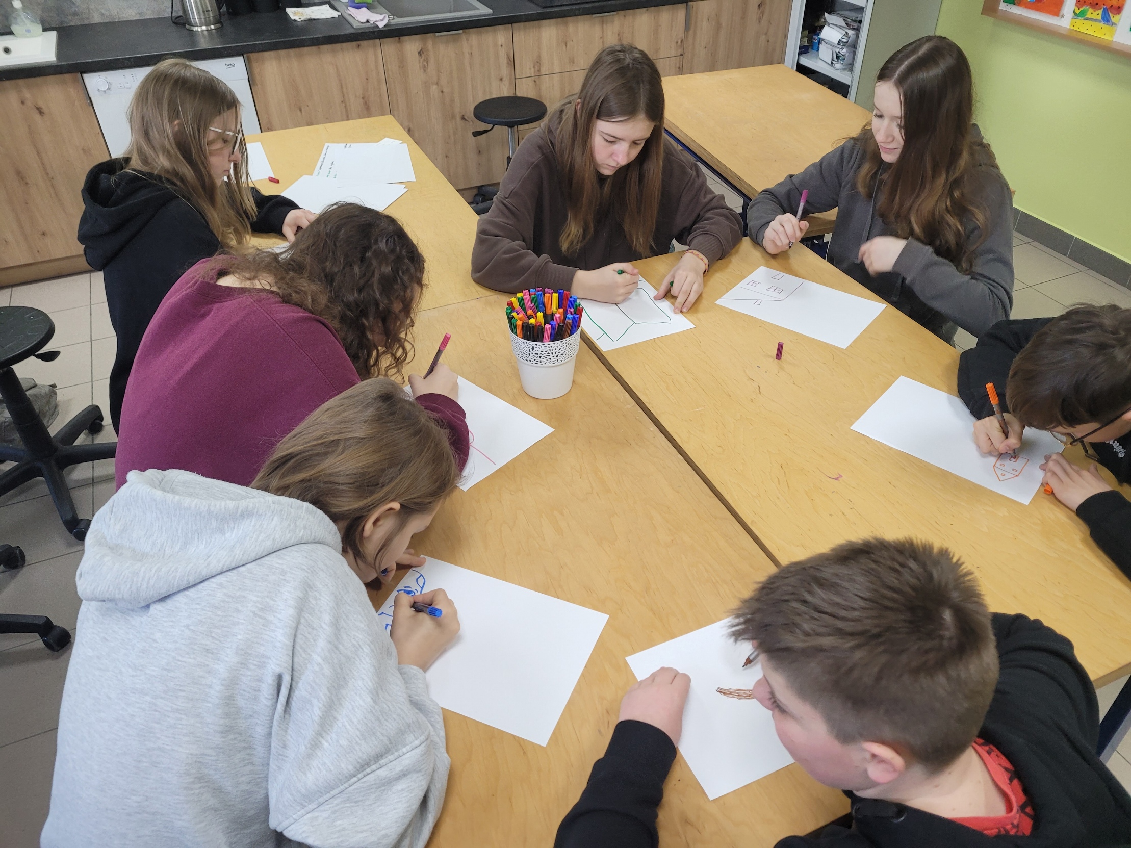 W sali lekcyjnej, grupa młodzieży siedzi w ławkach w dwóch rzędach, pochylona nad rozłożonymi na stole kartkami papieru.